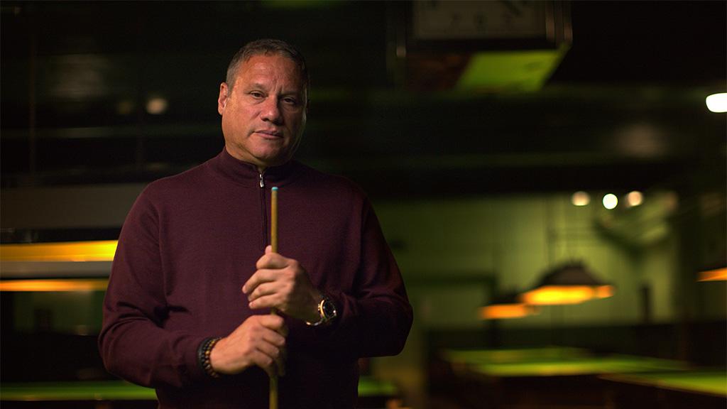 An older man holding a pool stick with a serious look on his face. Behind him is a blurred dark green and gold background of pool tables and lights