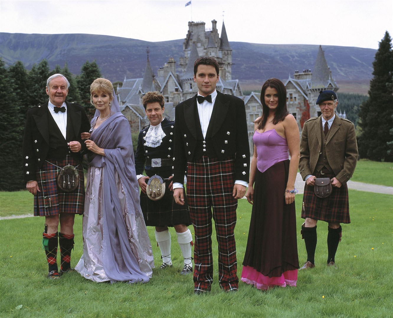 Six white people standing in a row in a field of grass, 3 older with white hair, and 3 younger with brown hair. All of which dressed for a fancy occasion