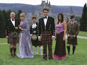 Six white people standing in a row in a field of grass, 3 older with white hair, and 3 younger with brown hair. All of which dressed for a fancy occasion