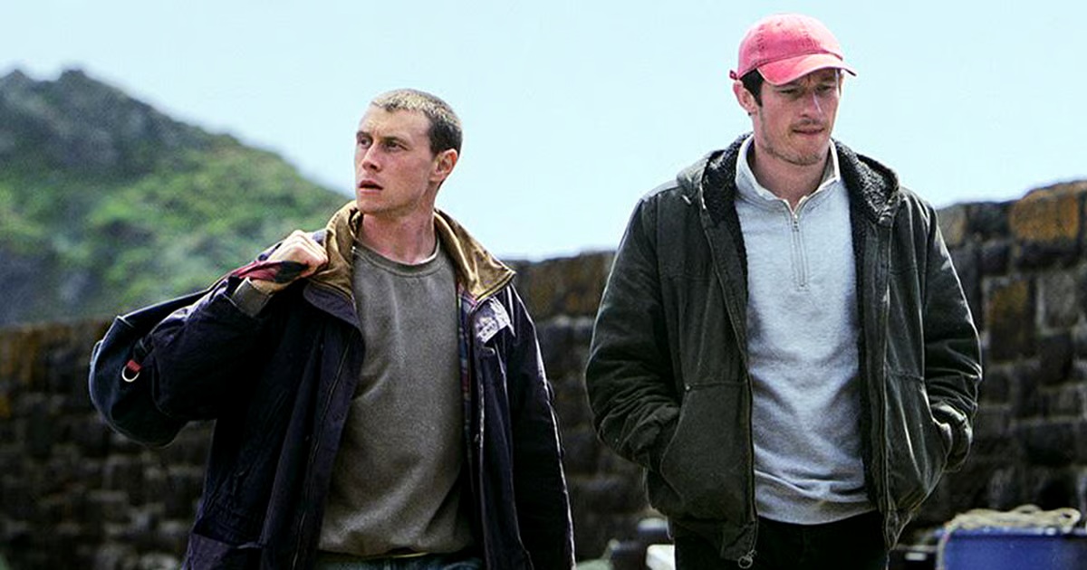 Two early 30's white men walking next to eachother away from hills in the background, both of which wearing a dark jacket. One looking down, the other (holding a bag) looking away