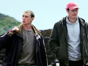 Two early 30's white men walking next to eachother away from hills in the background, both of which wearing a dark jacket. One looking down, the other (holding a bag) looking away