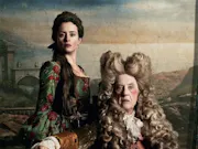 A late 30's white woman with dark hair standing next to a seated early-60s white man in front of a sunset backdrop. Both of which are in 18th century England attire, with the man wearing a large white wig