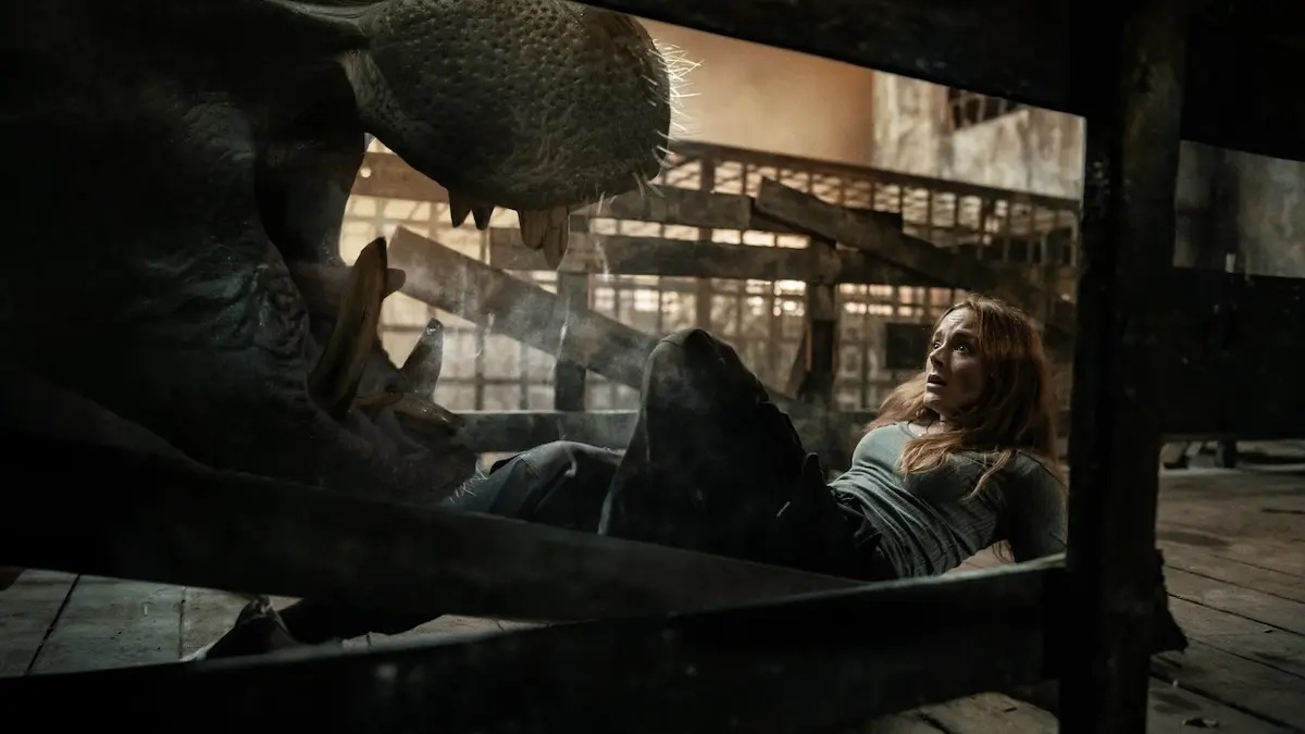 A mid-20's red-haired white woman on the floor with a scared look on her face, facing the open mouth of a hippo