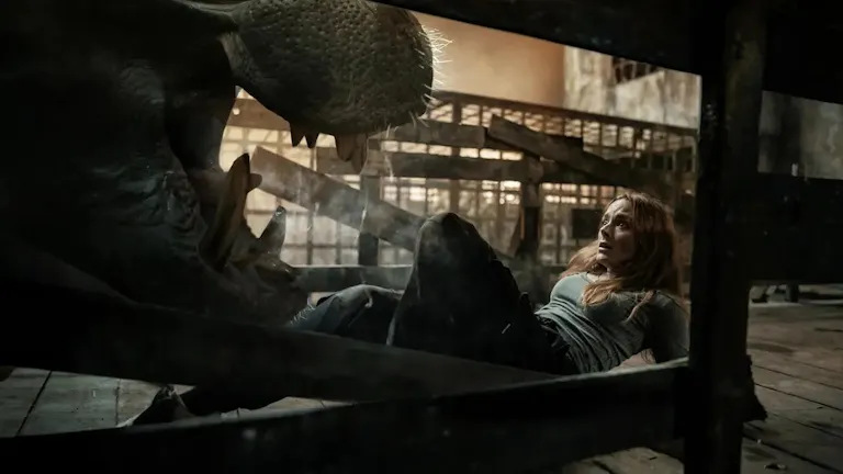 A mid-20's red-haired white woman on the floor with a scared look on her face, facing the open mouth of a hippo