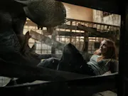 A mid-20's red-haired white woman on the floor with a scared look on her face, facing the open mouth of a hippo