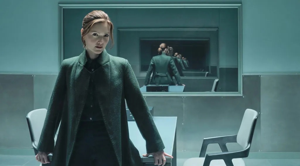 An early-30's white woman with red hair standing confidently in an interrogation room looking right at the camera. Behind her is the reflection of her back reflected many times in the one-way mirror
