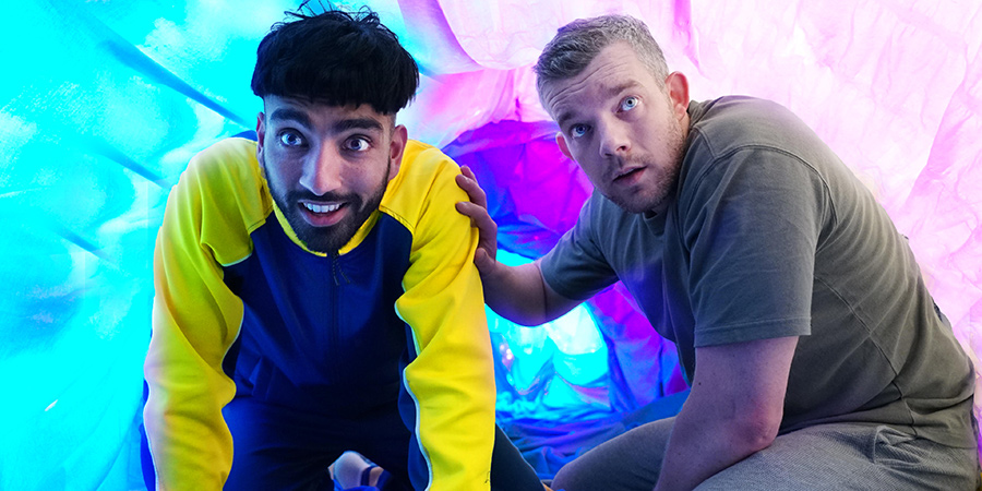 An olive skinned young boy with a bowl cut crouched next to a white middle aged man, both in front of a blue and purple fluorescent background