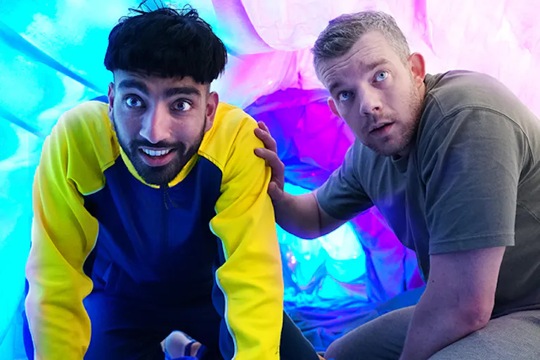 An olive skinned young boy with a bowl cut crouched next to a white middle aged man, both in front of a blue and purple fluorescent background