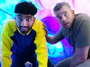 An olive skinned young boy with a bowl cut crouched next to a white middle aged man, both in front of a blue and purple fluorescent background