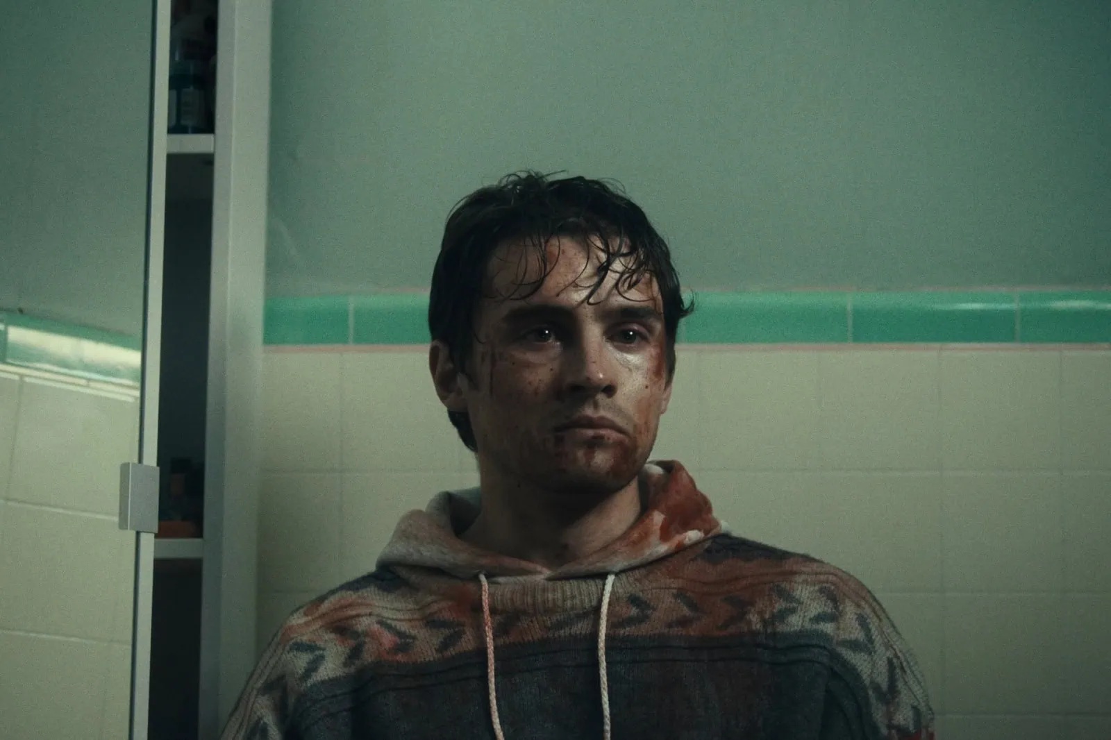 A young dark-haired white man with watery eyes and covered in blood, sitting against bathroom tiles with a blank stare