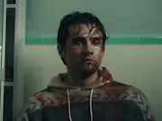 A young dark-haired white man with watery eyes and covered in blood, sitting against bathroom tiles with a blank stare
