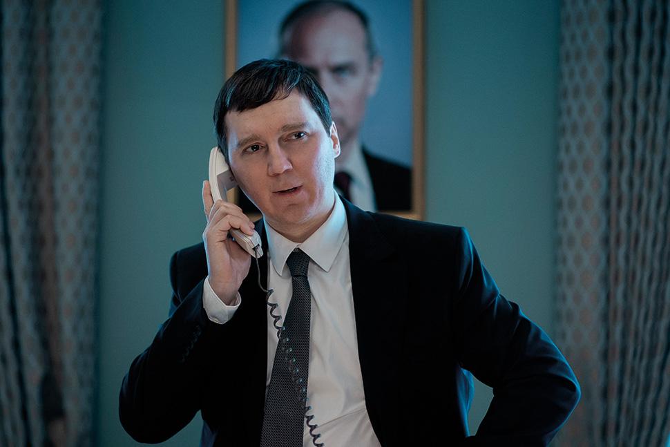 A white man with short dark hair, wearing a black suit inside of a presidential building. He is holding a landline phone to his ear and standing in front of a framed wall picture of another white man in a suit
