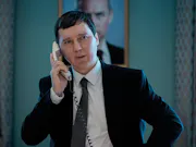 A white man with short dark hair, wearing a black suit inside of a presidential building. He is holding a landline phone to his ear and standing in front of a framed wall picture of another white man in a suit