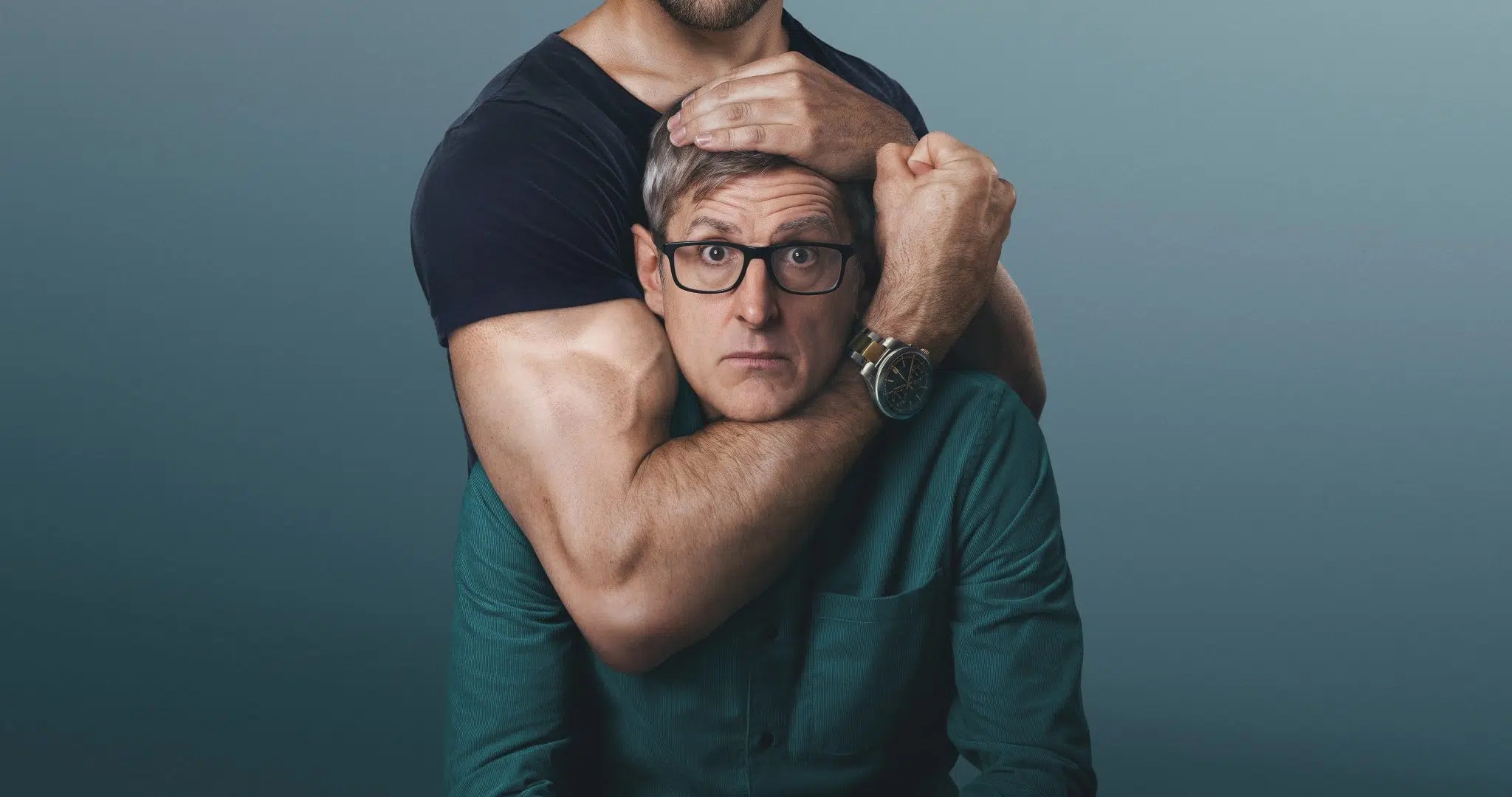A middle-aged white man wearing glasses, being held in a headlock position by muscular olive skinned man, who's face is out of frame