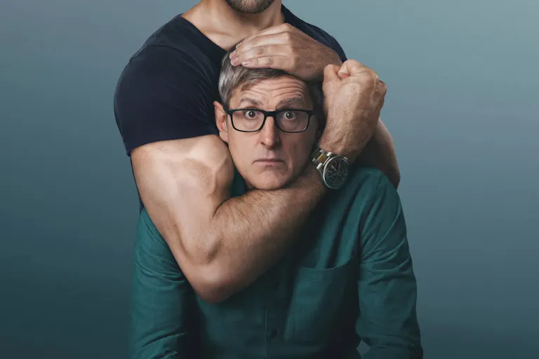 A middle-aged white man wearing glasses, being held in a headlock position by muscular olive skinned man, who's face is out of frame