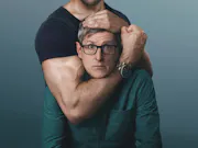 A middle-aged white man wearing glasses, being held in a headlock position by muscular olive skinned man, who's face is out of frame