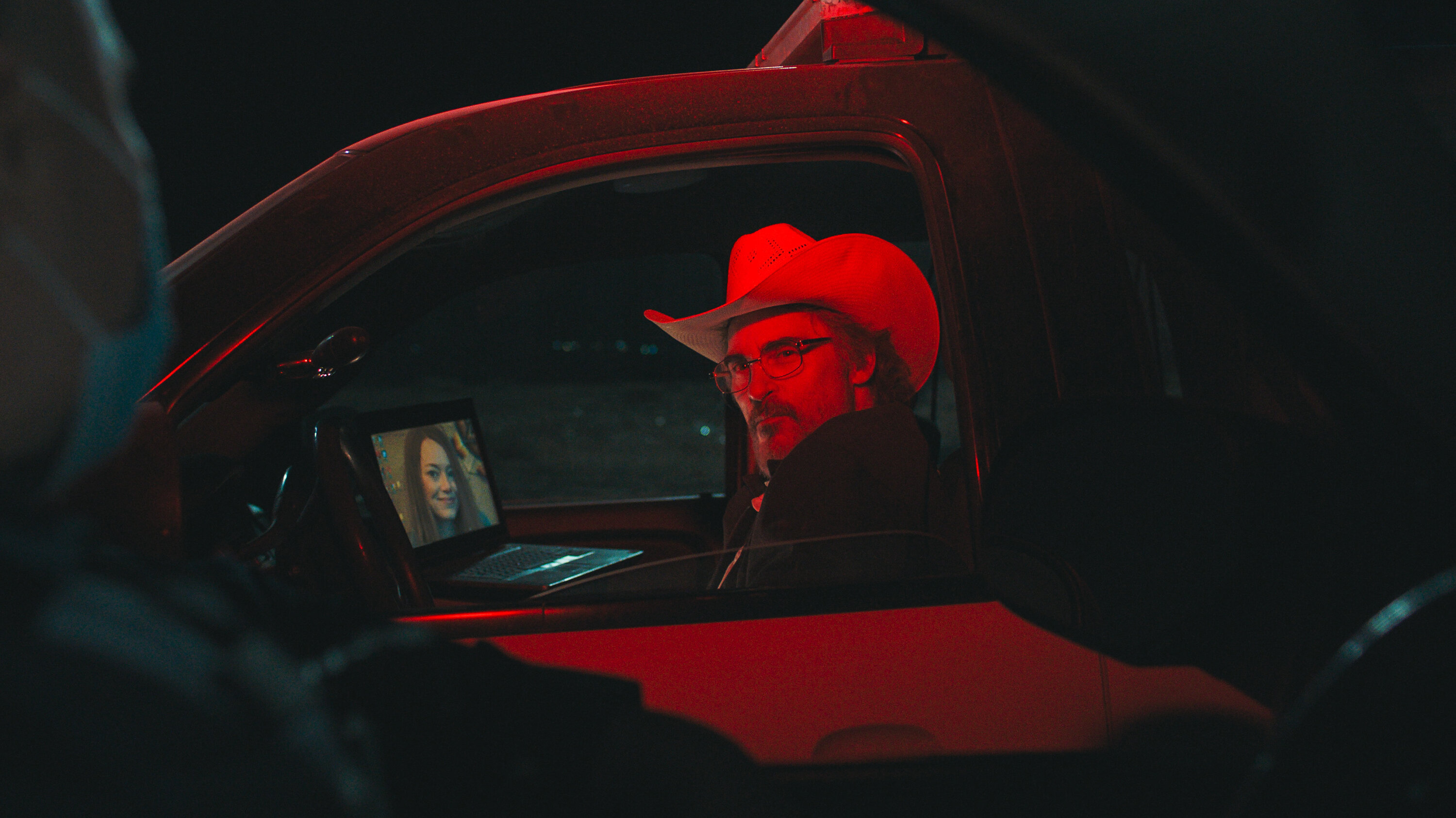 A middle-aged white man with a cowboy hat, with a smug expression while looking out the window of his truck. Shining on him is a light which makes the whole image red, Resting on the steering wheel is an open laptop with a middle-aged long-haired smiling woman as the screensaver