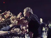 A white man in a black hoodie facing dozens of hands stretched out towards him