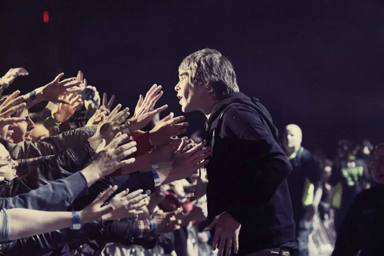 A white man in a black hoodie facing dozens of hands stretched out towards him