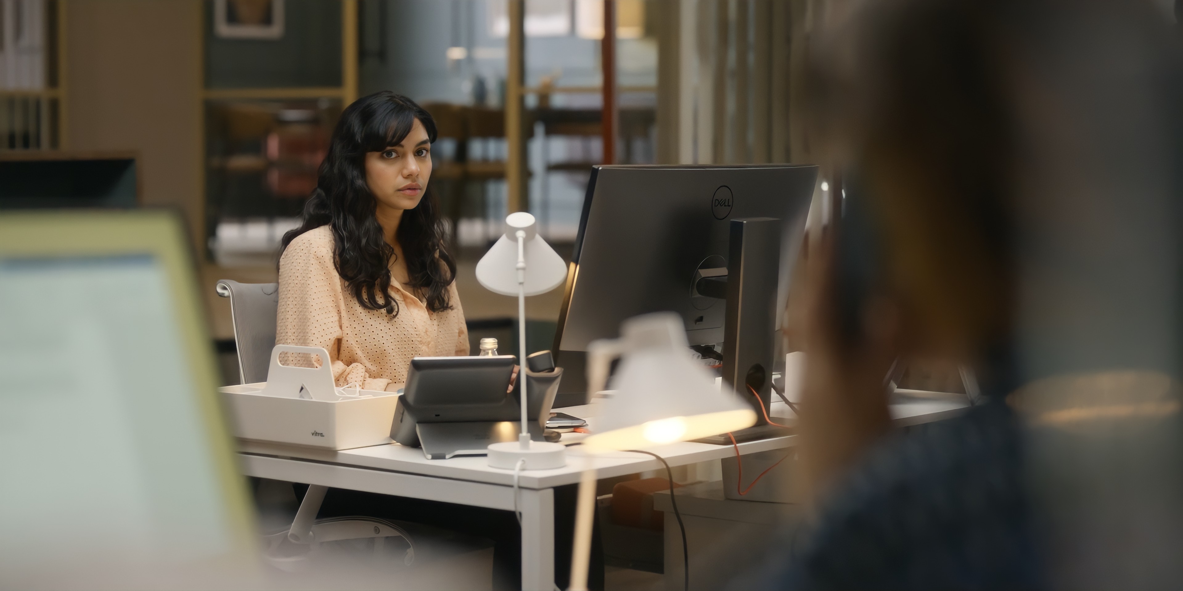 A young south asian woman with long black hair sitting at a work desk, looking worriedly at the blurred person in the foreground
