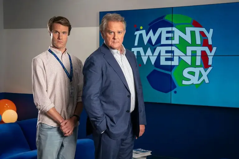 A middle aged white man wearing a shirt and blue lanyard, next to an older white man wearing a suit jacket and shirt, both in front of a sign on the wall which reads 'Twenty Twenty Six'