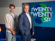 A middle aged white man wearing a shirt and blue lanyard, next to an older white man wearing a suit jacket and shirt, both in front of a sign on the wall which reads 'Twenty Twenty Six'