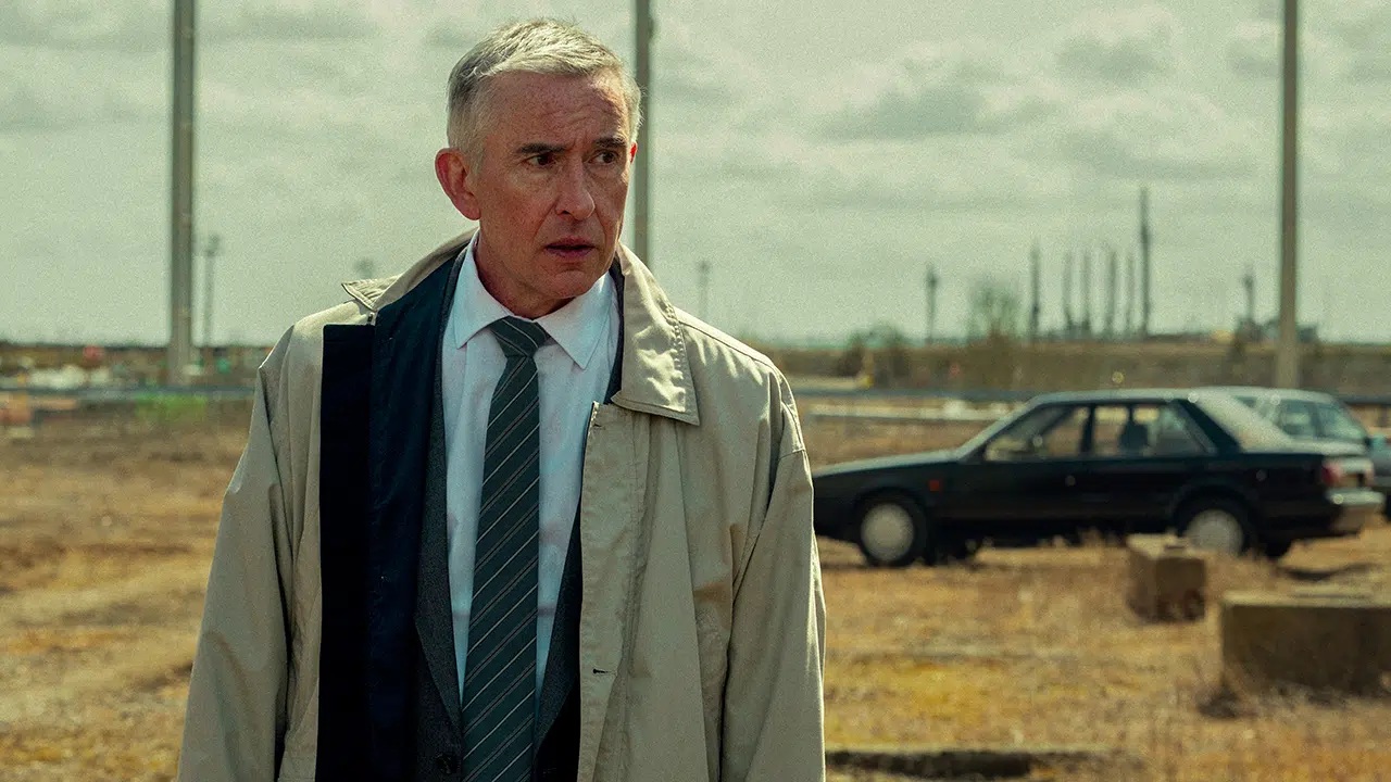 A grey-haired older white man wearing a suit and detective jacket, standing in a field of brown grass, with abandoned vehicles in the background