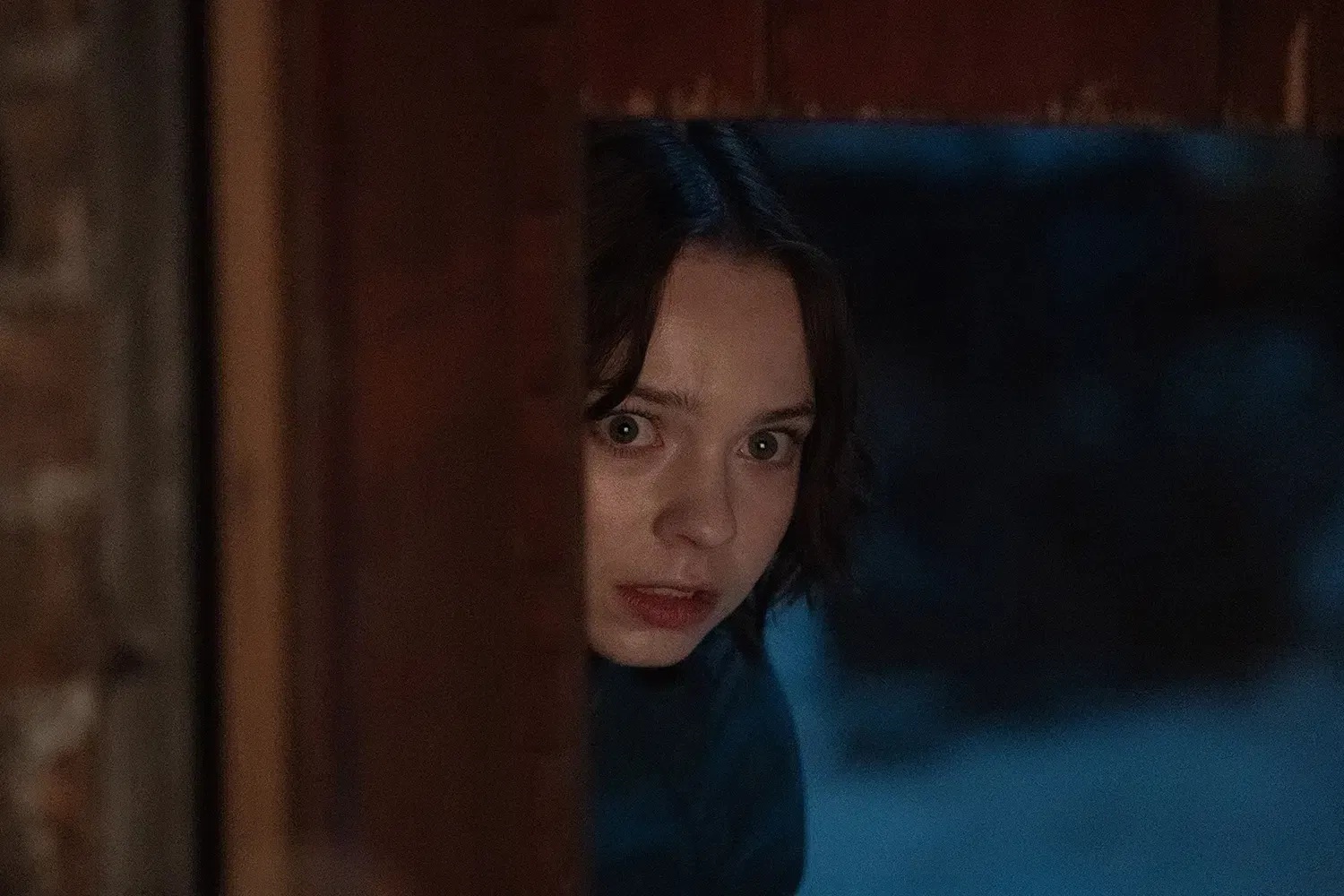 A young brown-haired caucasian woman peeking from around a corner, with a scared face