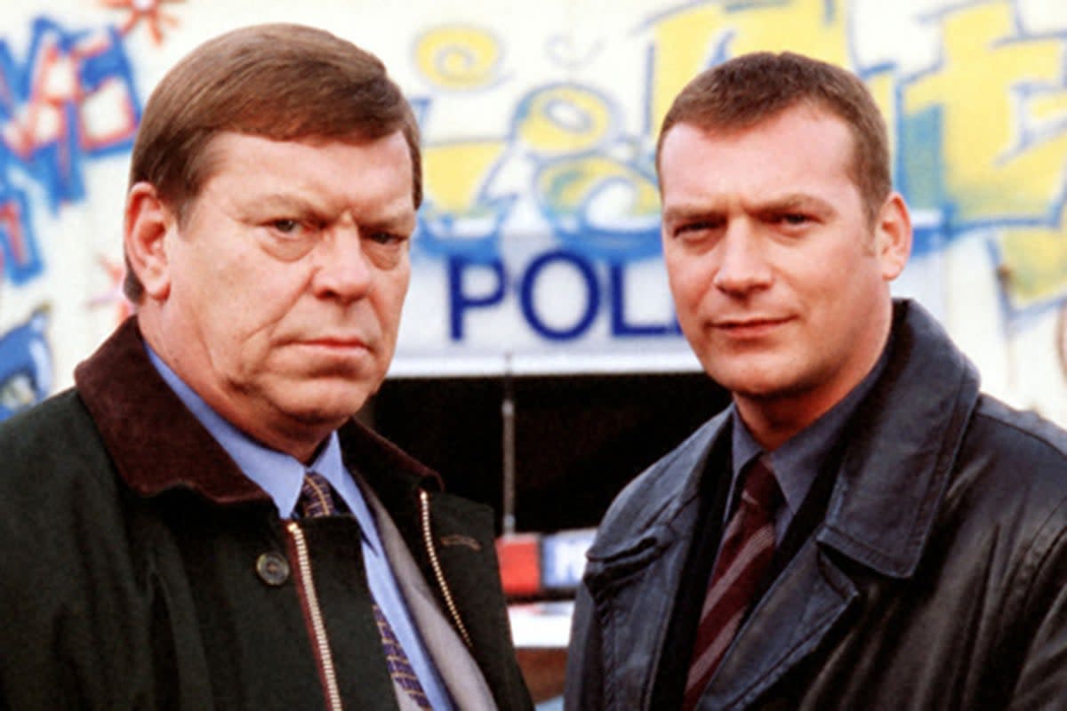 Two white men in shirts, ties and jackets with stern faces, standing in front of a police station