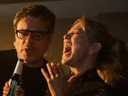 A mid-30s white man with glasses next to a mid-30s blonde white woman with her hair tied up. Both are singing something; the woman with a widened mouth and eyes closed, and the man singing into the bottle the woman is holding