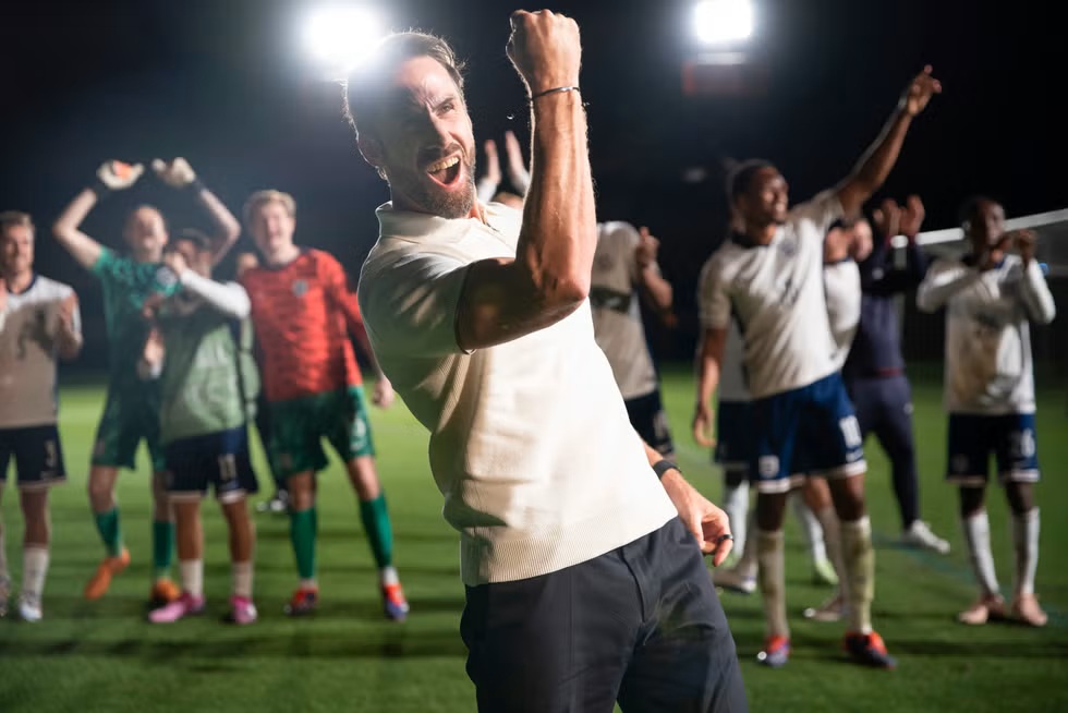 A mid-30s white man cheering with his mouth open and first upwards. In the background, numerous men in different coloured football kits, also cheering