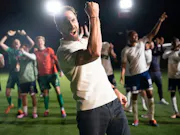 A mid-30s white man cheering with his mouth open and first upwards. In the background, numerous men in different coloured football kits, also cheering