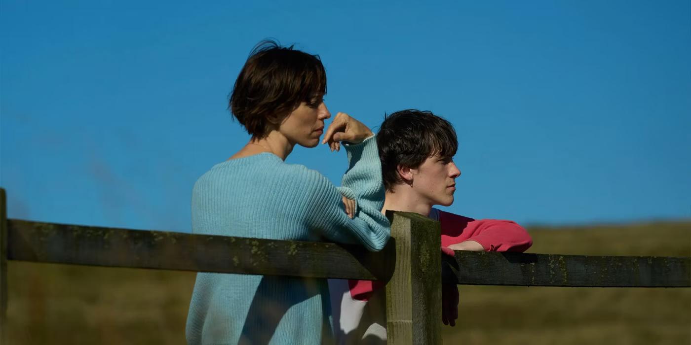 A middle aged dark-haired white woman and a 20-something dark-haired white boy leaning on a fence in the middle of a field, looking off into the distance. Behind them you can see the blue sky