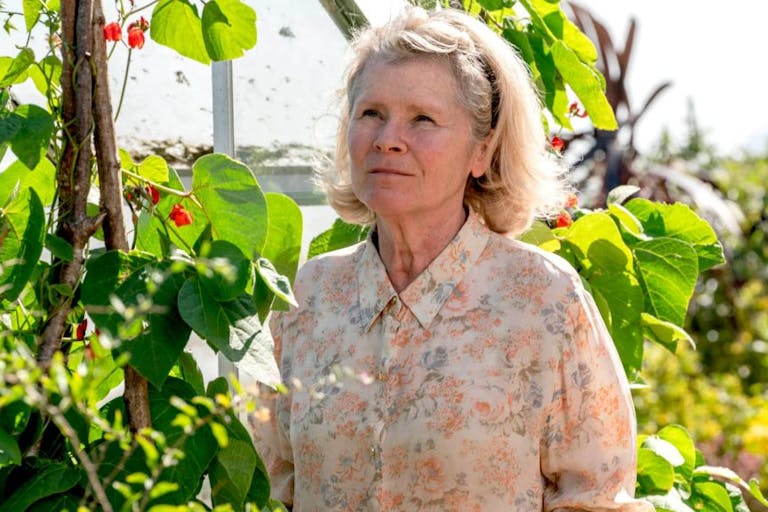 An older white woman in a floral shirt stands in a sunny garden