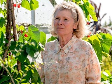 An older white woman in a floral shirt stands in a sunny garden 