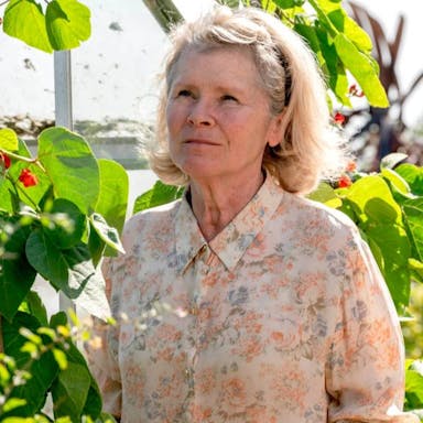 An older white woman in a floral shirt stands in a sunny garden 