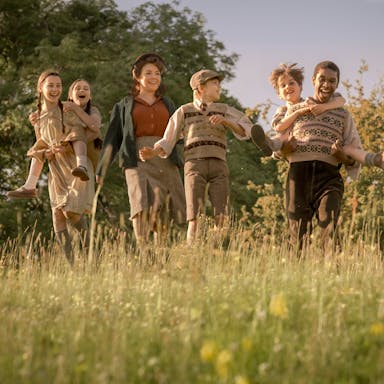 A mix of seven children and adults laughing and playing piggyback in a sunny field, wearing period clothing 