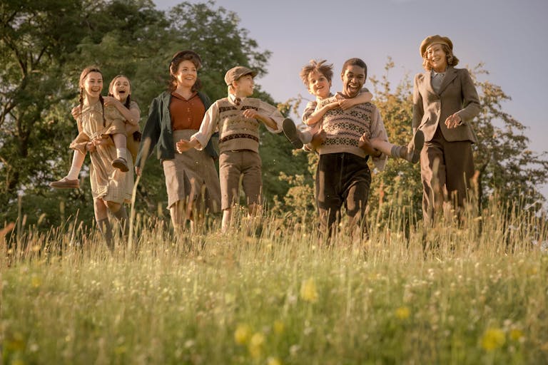 A mix of seven children and adults laughing and playing piggyback in a sunny field, wearing period clothing