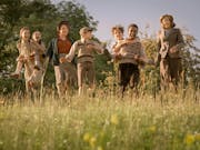 A mix of seven children and adults laughing and playing piggyback in a sunny field, wearing period clothing