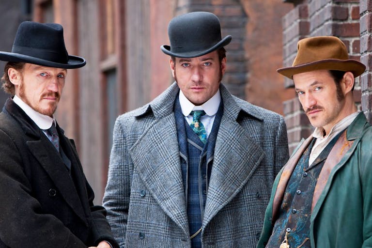 Three men in smart period business clothes and hats staring seriously at the audience