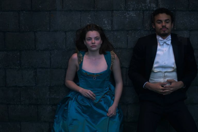 A young white woman in an elegant blue corset dress and young black man in a smart black and white tux, lying on a stone floor beside each other