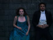 A young white woman in an elegant blue corset dress and young black man in a smart black and white tux, lying on a stone floor beside each other