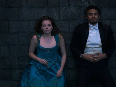 A young white woman in an elegant blue corset dress and young black man in a smart black and white tux, lying on a stone floor beside each other