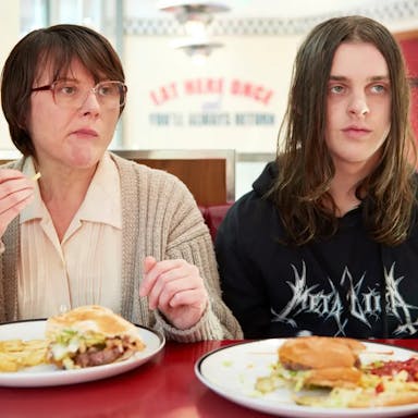 An older white woman and young white male teen with long hair and Metallica hoodie sitting in a diner eating burger and chips