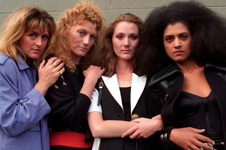 Three white women and one Black woman stand with their arms linked and around each other