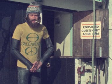 Dated image of a bearded white man in a wetsuit, woollen hat, and yellow 'Greenpeace' shirt with a cigarette in his mouth, on board a boat