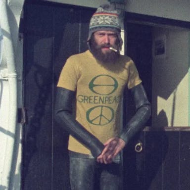 Dated image of a bearded white man in a wetsuit, woollen hat, and yellow 'Greenpeace' shirt with a cigarette in his mouth, on board a boat