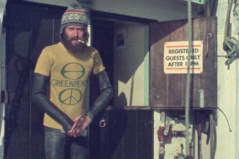 Dated image of a bearded white man in a wetsuit, woollen hat, and yellow 'Greenpeace' shirt with a cigarette in his mouth, on board a boat