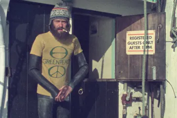 Dated image of a bearded white man in a wetsuit, woollen hat, and yellow 'Greenpeace' shirt with a cigarette in his mouth, on board a boat