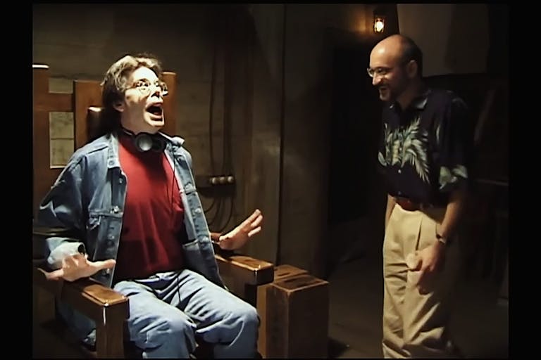 Behind the scene shot of two white men, one sat in a chair fake screaming and the other standing laughing